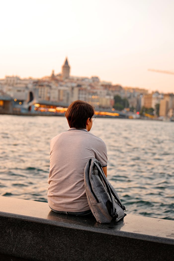 about-us Person sits pensively by the water with cityscape backdrop at sunset, evoking reflection.